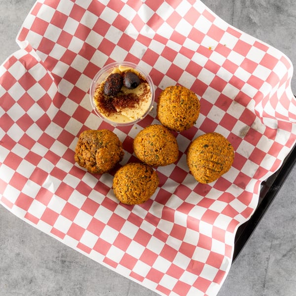 Five falafel balls and a small container of hummus with olives on red and white checkered paper.