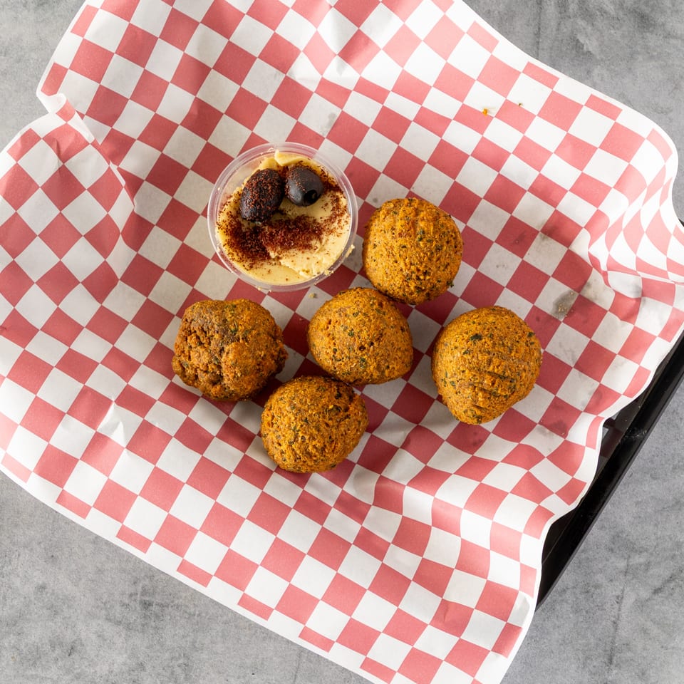 Five falafel balls and a small container of hummus with olives on red and white checkered paper.