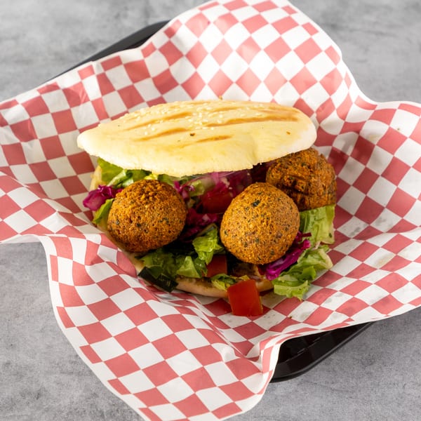 A falafel pita sandwich with lettuce, red cabbage, and tomatoes, served in a red and white checkered paper.