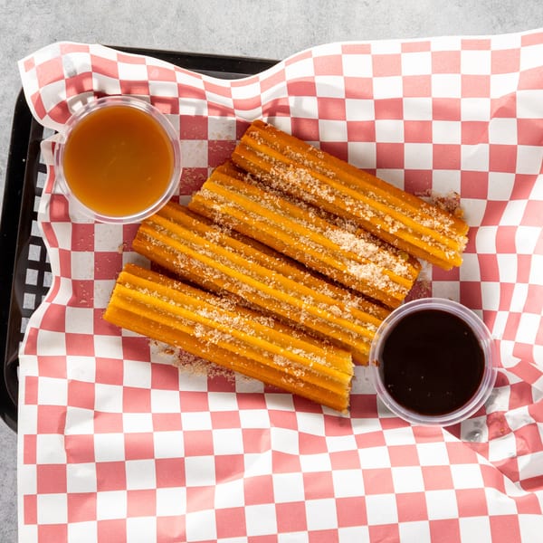 Churros sprinkled with sugar, served in a basket with red and white checkered paper, with caramel and chocolate dipping sauces.