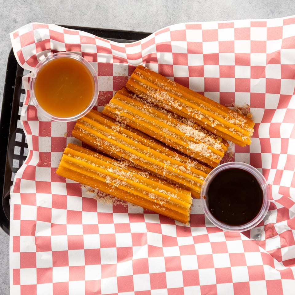 Churros sprinkled with sugar, served in a basket with red and white checkered paper, with caramel and chocolate dipping sauces.