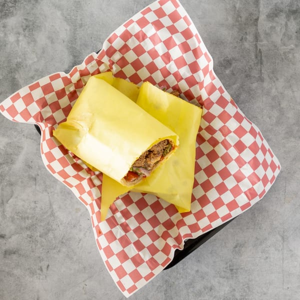 Two yellow-wrapped sandwiches with meat and vegetables, served in a red and white checkered paper-lined basket.